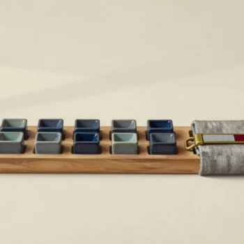 Two rows of small ceramic vessels on a wooden tray beside folded woven cloth with a brass clasp.