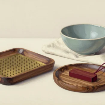A perforated brass insert in a rectangular wooden tray beside a vermilion wooden piece on a round tray and a sage bowl.