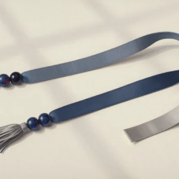 A long looped slate-blue ribbon with ceramic beads and tassels at its ends beside a short gray ribbon.