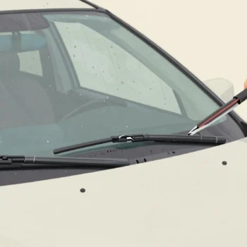 A hand fitting a new wiper blade to a parked car in the rain, the old cracked blade resting on the windshield