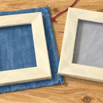 Two ivory frames lie on a wooden table, one over blue woven cloth and one with a plain gray inset.