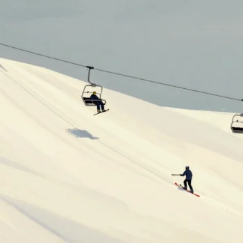 A skier riding a chairlift while a groomer prepares the slope below