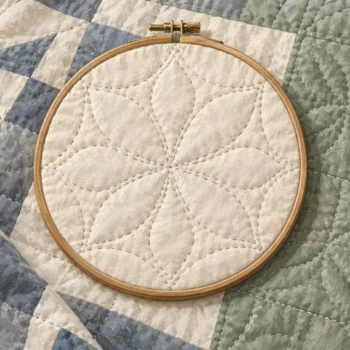 A quilting hoop holds a quilt with adjoining pale and sage fabric sections