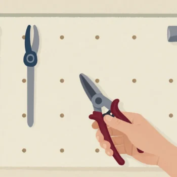 A hand hanging pruning shears on a shed pegboard where each tool has a painted outline behind it