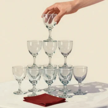 A hand lifts the top glass from a pyramid of stacked glasses, a red napkin at its base.