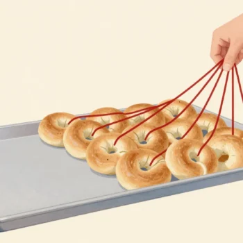 A hand threading loose bagels from a tray onto one long red string at a bakery stall