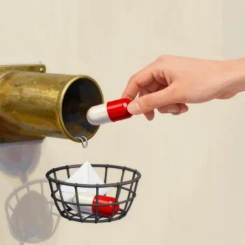 A hand sending a red capsule into a brass pneumatic tube as a reply capsule drops into the basket below