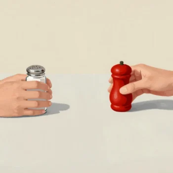 Two hands across a table, one holding salt and one holding a red pepper shaker, each reaching for the other's.