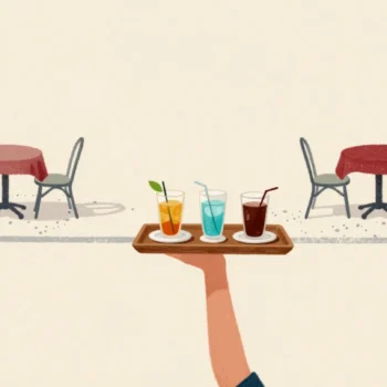 A hand carrying three drinks and a plate on one wooden tray across a cafe terrace, worn gravel paths behind.