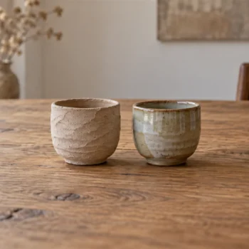 An unfinished clay cup sits beside a smooth finished cup on a potter's workbench.