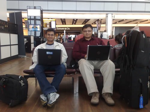 Suprotim and Pinal at London Airport Two men with laptops sitting on bench in airport.