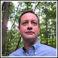 A man in a blue shirt stands in front of a forest.