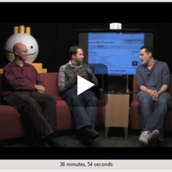 Two men talk with a large silver coin figure.