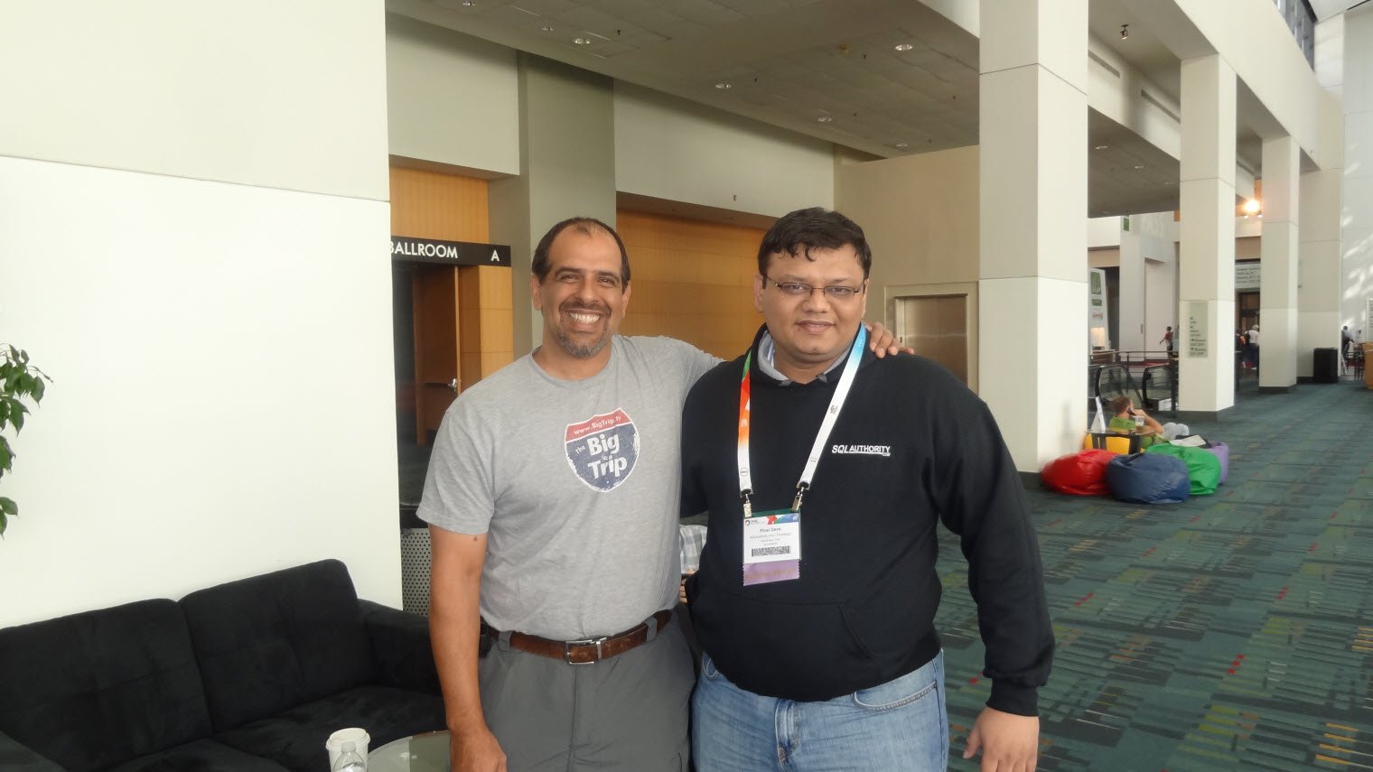 Two men standing indoors, one wearing 'Big Trip' T-shirt.